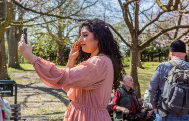 Danimarka, Kopenhag 'daki Bispegjerg mezarlığında kiraz ağacı bahar çiçeği açarken selfie çeken kadın.