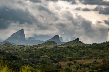 Pedra do Bispo 'nun çarpıcı granit tepeleri Conda, Angola' nın gür, tropikal manzarası üzerinde kasvetli bulutların arasından yükseliyor. Afrika jeolojisi ve vahşi doğasının büyüleyici bir görüntüsü, seyahat, doğa ve atmosferik temalar için mükemmeldir..