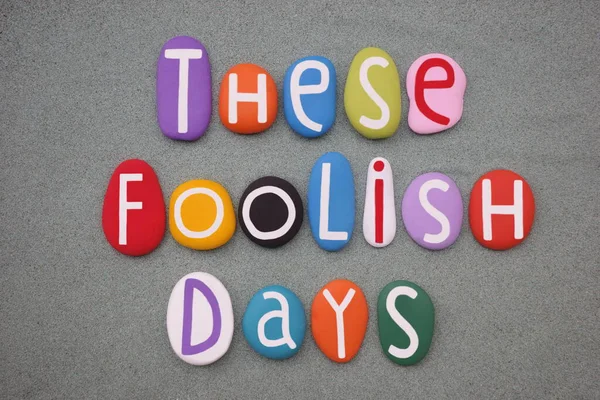 stock image These foolish days, creative slogan composed with multi colored stone letter over green sand