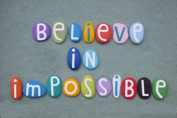 Believe in impossible, creative motivational phrase composed with hand painted multi colored stone letters over green sand