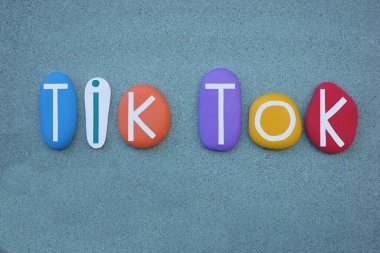 Tik, tok, sound of a clock making a regular clicking sound, creative text composed with multi colored stone letters over green sand