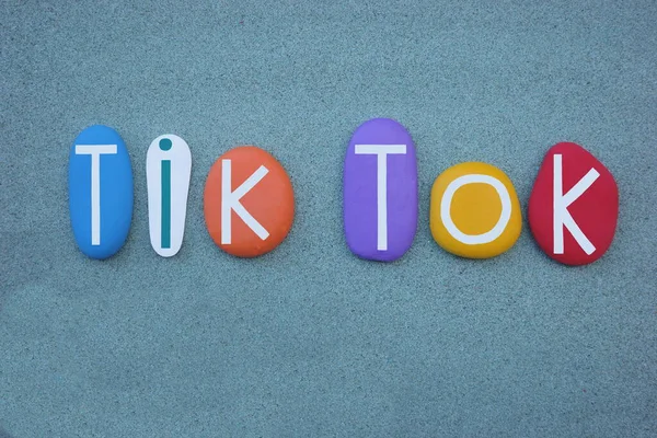 Tik, tok, sound of a clock making a regular clicking sound, creative text composed with multi colored stone letters over green sand