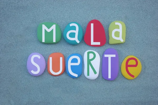 Mala suerte, Bad luck in spanish language composed with hand painted multi colored stone letters over green sand