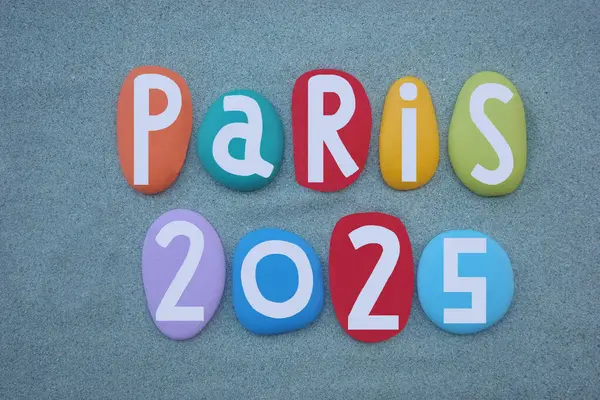 stock image Paris 2025, creative logo composed with hand painted multi colored stone letters over green sand