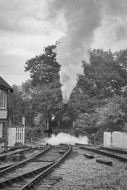 Küçük bir hafif buhar lokomotifi kameraya doğru ilerlerken içinden duman ve buhar çıkan bir istasyona doğru ilerliyor.