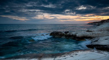 Kayalık bir sahilde dramatik bir karamsar gökyüzü ile güzel bir gün batımı. Paphos Kıbrıs