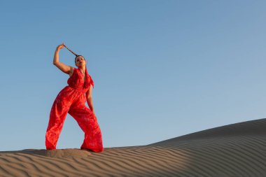Çölde kumullarda dans eden kırmızı giysili çekici bir kadın mavi gökyüzüne karşı..