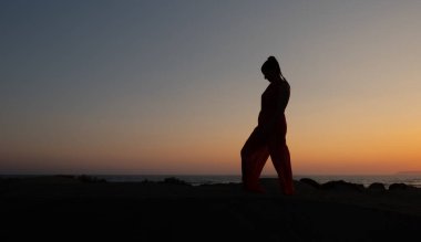 Gün batımında kumlu bir sahilde dans eden bir kadının silueti. Beyaz kadın sahilde poz veriyor..