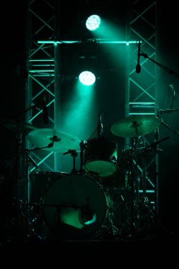 Rock festivalinde konser sahnesi, müzik aletleri davul siluetleri, renkli arka plan.