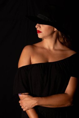 Siyah arka planda el ele tutuşan siyah şapkalı beyaz bir kadının portresi.