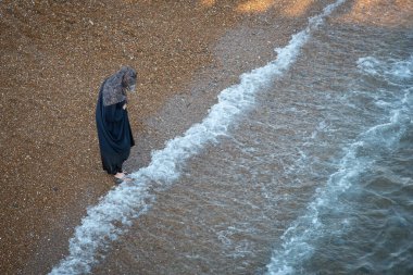 Brighton, Birleşik Krallık, 10 Haziran 2023: Geleneksel siyah elbiseli Arap kadın sahilde yürüyor.