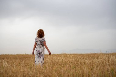 Gündelik giysiler içinde buğday tarlasında yürüyen yalnız bir genç kadın. Bulutlu gökyüzü