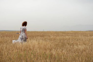 Gündelik giysiler içinde buğday tarlasında yürüyen yalnız bir genç kadın. Bulutlu gökyüzü