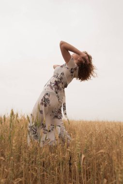 Çiçekli elbiseli mutlu kadın altın buğdayın ortasında duruyor, huzurlu manzaranın tadını çıkarıyor.