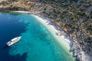 Turistlerle dolu turkuaz bir plajın İyon Denizi 'ndeki Kefalonya Yunan adasının yakınlarında demirleyen bir tur teknesinin insansız hava görüntüsü.