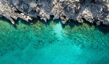 Turkuaz deniz suyunun hava aracı görüntüsü güneşli bir yaz gününde kayalık bir sahil şeridiyle buluşuyor.