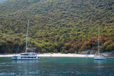 Issız bir tropikal plajın yakınında demirli lüks yatlar. Kumsalın yukarısındaki orman. Kefalonia Adası, Yunanistan. İyon denizi, Yunan adaları