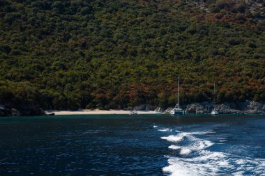 Sürat teknesi, yakınlarda yelkenli tekneleriyle el değmemiş tropikal bir sahilden uzaklaşırken köpüklü bir şekilde kalkıyor. Yunanistan 'ın Kefalonia adası