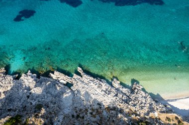 Turkuaz deniz suyunun hava aracı görüntüsü güneşli bir yaz gününde kayalık bir sahil şeridiyle buluşuyor.