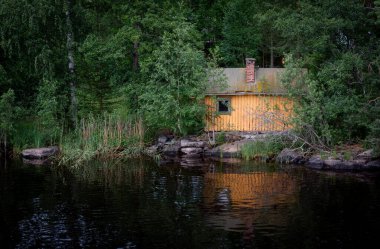 Ormandaki küçük ahşap sauna evi gün batımında sakin göl suyuna yansıyor.