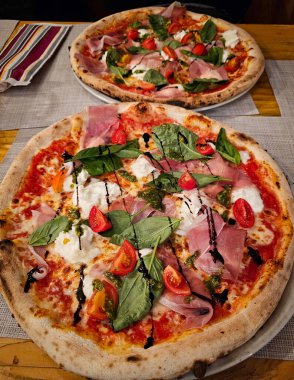 Ahşap bir restoran masasında prosciutto, burrata peyniri, pesto ve vişneli domatesli iki taze yuvarlak pizza servis edilir.
