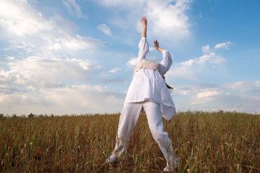 Açık havada mavi bulutlu gökyüzüne karşı dans eden beyaz elbiseli genç bir kadın. Dışarıda tasasız bir kadın. Doğada aktif olan insanlar