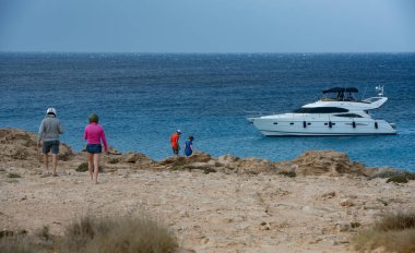 Ayia Napa, Kıbrıs, 13 Mayıs 2024: Kayalık bir kıyı şeridi boyunca yürüyen turistler, mavi okyanusta demirlemiş lüks bir yata bakıyorlar. Cape Greko Ayia Napa Kıbrıs