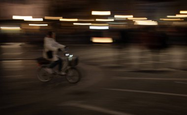 Geceleri şehirde bisiklet süren genç bir kadın bulanık ışıklarla hız ve hareket hissi yaratıyor.