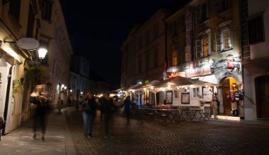 Ljubljana, Slovenya, 30 Ekim 2024: Avrupa 'nın başkenti Slovenya' nın başkenti Ljubljana 'da gece yarısı açık oturma yerleri aydınlatılmış bir şekilde parke taşı sokağından geçen turistler görülüyor