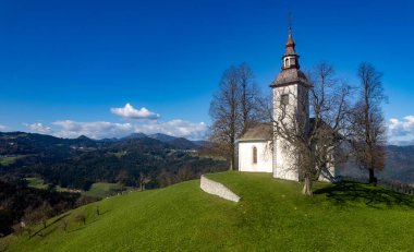 Slovenia 'da yeşil bir tepenin üzerinde duran Aziz Tomaz kilisesinin manzarası, sonbahar doğası ve ufukta bir dağ sırası ile çevrili. Aziz Tomas Köyü Slovenya