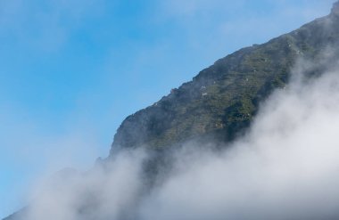 Beyaz bulutlar yükseliyor ve açık mavi gökyüzünün altında yeşil bitki örtüsüyle bir dağ yamacını örtüyor. Julian Alps dağ zirveleri, Slovenya