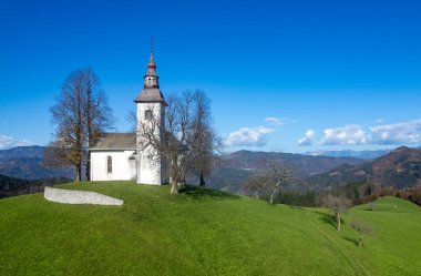 Slovenia 'da yeşil bir tepenin üzerinde duran Aziz Tomaz kilisesinin manzarası, sonbahar doğası ve ufukta bir dağ sırası ile çevrili. Aziz Tomas Köyü Slovenya