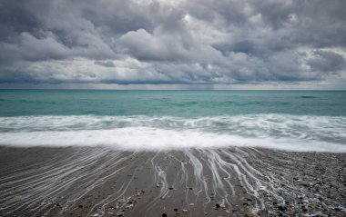 Turkuaz bir okyanusun üzerinde dramatik bulutlar çakıl taşlı bir plaja çarpan dalgalar güzel ve karamsar bir deniz manzarası yaratıyor.