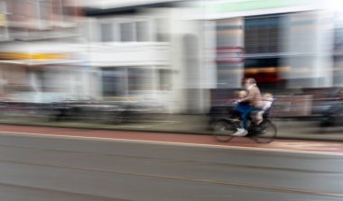 Babam bisikletiyle iki çocuk taşıyor, şehir manzarasına karşı hareket bulanıklığı yaratıyor.