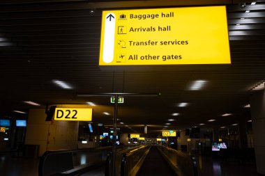 Aydınlatılmış sarı havaalanı tabelaları yolcuları geliş, bagaj teslim, nakil hizmetleri ve modern bir havaalanı terminalinin kapıları için rehberlik ediyor.