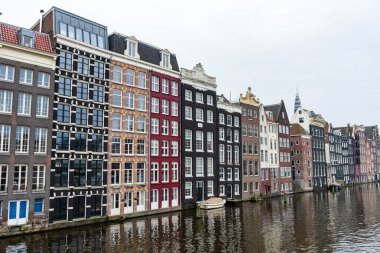 Hollanda, Amsterdam 'da su yüzeyine yansıyan demirlemiş teknelerle kanal boyunca uzanan renkli dar binalar dizisi.