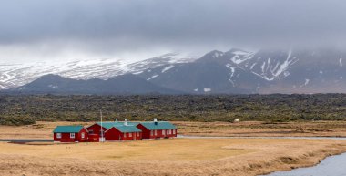 Geniş lav alanı ve bulutlu bir gökyüzünün altında karla kaplı dağlarla geleneksel İzlanda kırmızı evlerinin manzarası. İzlanda yolculuk macerası