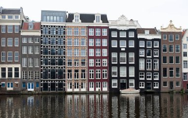 Hollanda, Amsterdam 'da bir kanala yansıyan renkli dar binalar dizisi, bulutlu bir günde, önünde demirlemiş küçük bir tekne.