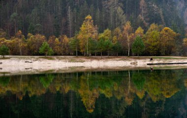 Renkli sonbahar ağaçları sakin bir gölün yüzeyine yansıyor. Fotoğrafçı, Jasna Slovenya Gölü 'ndeki ahşap iskelede duruyor.