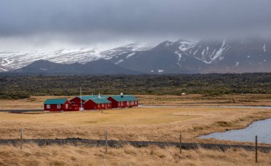 Geniş lav alanı ve bulutlu bir gökyüzünün altında karla kaplı dağlarla geleneksel İzlanda kırmızı evlerinin manzarası. İzlanda yolculuk macerası