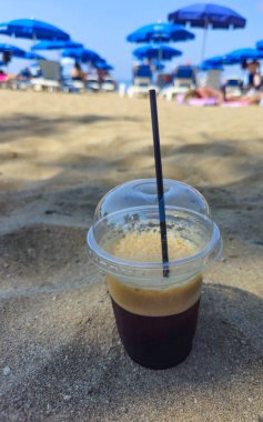 Kumlu bir sahilde soğuk, serinletici bir buzlu kahve. Freddo espresso yaz mevsiminde ferahlatıcıdır. Tatil köyündeki plaj şemsiyeleri.