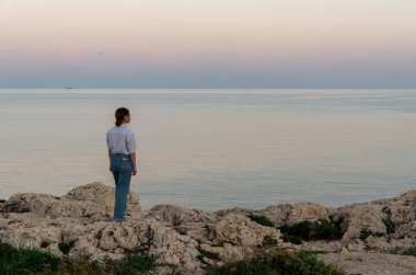 Genç bir kadın sakin bir gün batımında sakin okyanusa bakarak kayalık bir sahilde duruyor.