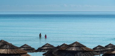 Güneşli bir günde plaj şemsiyeleri gölgeli sakin turkuaz denizde ferahlatıcı bir yüzüşün keyfini çıkaran çift.
