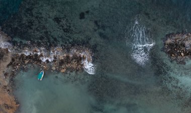Küçük bir balıkçı teknesinin sakin hava görüntüsü kayalık bir sahil şeridine demirlemiş, dalgalar yavaşça çarpıyor, sakin bir sahil manzarası yaratıyor.
