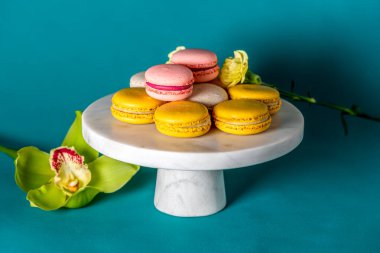 French macaron cakes on a marbel white cake stand against blue backdrop close up. Yellow white, pink macarons. Orchid and sage.