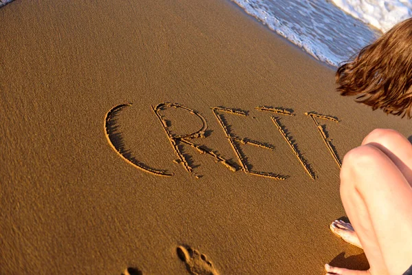 The word crete written in the sand at the water\'s edge. A woman is writing word Crete on sea sand.