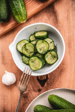Ahşap bir masada seramik beyaz bir kasede organik salatalıklar dilimler. Taze sebze ve dereotu, ahşap dokuma masasında sarımsak. Salata..