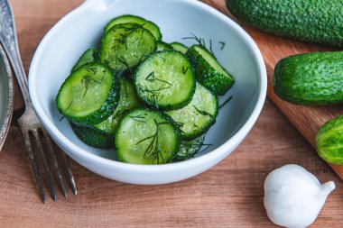 Ahşap bir masada seramik beyaz bir kasede organik salatalıklar dilimler. Taze sebze ve dereotu, ahşap dokuma masasında sarımsak. Salata..