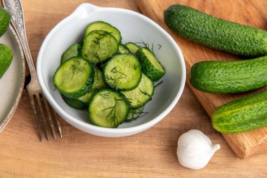 Ahşap bir masada seramik beyaz bir kasede organik salatalıklar dilimler. Taze sebze ve dereotu, ahşap dokuma masasında sarımsak. Salata..