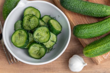 Ahşap bir masada seramik beyaz bir kasede organik salatalıklar dilimler. Taze sebze ve dereotu, ahşap dokuma masasında sarımsak. Salata..